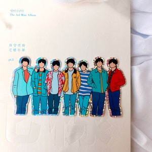 May include: A holographic sticker featuring a group of seven cartoon figures. The figures are all wearing different colored clothing and have black hair. The sticker is on a white background with the text "방탄소년단 The 3rd Mini Album 화양연화 pt.1" printed on it.