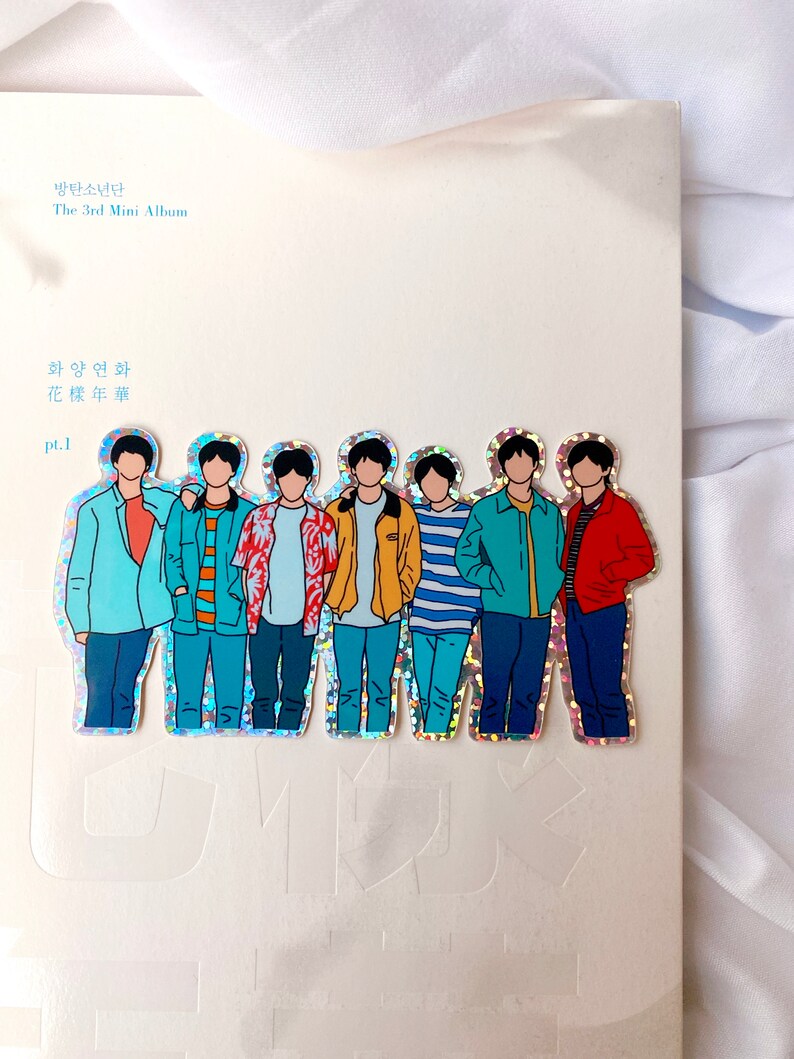 May include: A holographic sticker featuring a group of seven cartoon figures. The figures are all wearing different colored clothing and have black hair. The sticker is on a white background with the text "방탄소년단 The 3rd Mini Album 화양연화 pt.1" printed on it.