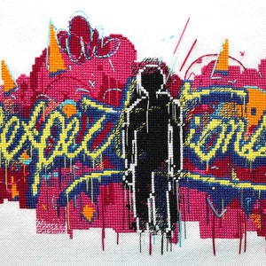 Può includere: Opera di ricamo a punto croce che raffigura una persona in piedi davanti a un muro di graffiti con le parole "Expect" e "More" in lettere gialle e blu.