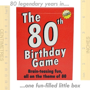 May include: A red box with the text "The 80th Birthday Game" in white. The box has a yellow star with the word "NEW" on it. The text "Brain-teasing fun, all on the theme of 80" is printed below the game title. The box is sitting on a wooden ruler with inch markings.