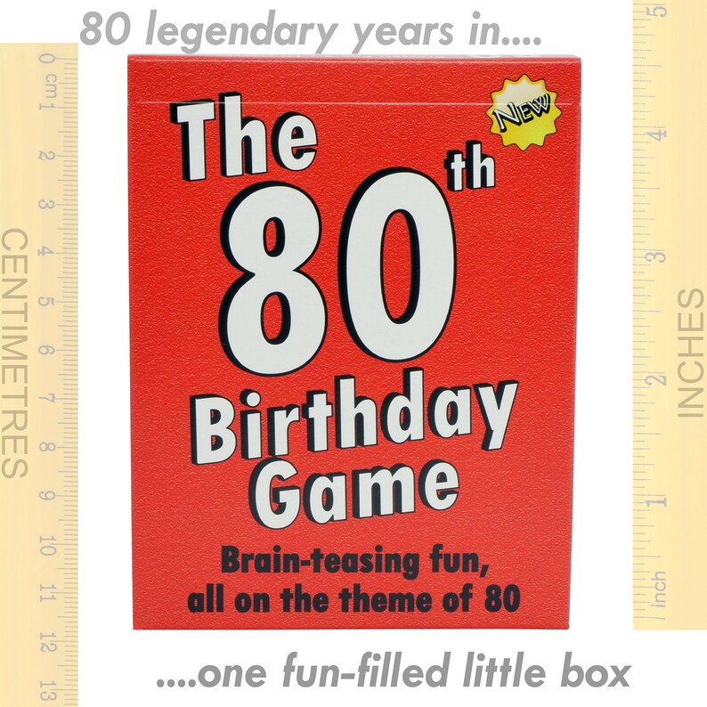 May include: A red box with the text "The 80th Birthday Game" in white. The box has a yellow star with the word "NEW" on it. The text "Brain-teasing fun, all on the theme of 80" is printed below the game title. The box is sitting on a wooden ruler with inch markings.