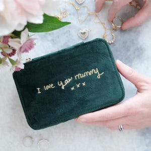 May include: A dark green velvet jewelry box with gold embroidered text reading "I love you mummy". The rectangular box is closed with a gold zip. Gold necklaces and rings are also visible in the image.