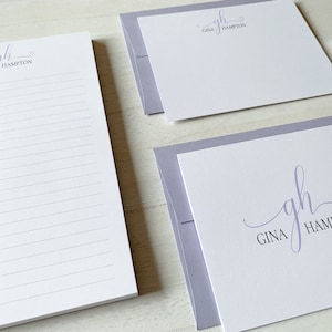 May include: A set of stationery featuring a light purple envelope with a white card inside. The card has a handwritten monogram "gh" with the name "Gina Hampton" printed below. The set also includes a lined notepad with the same monogram and name printed at the top.