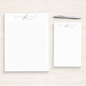 May include: Two white lined notepads with a simple cursive monogram design. The monogram is "jh" and the name "Judy Hamilton" is printed below the monogram. The notepads are on a light wood surface.