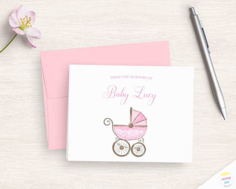 May include: A white card with pink text that reads "From the nursery of Baby Lucy". A pink and white baby carriage with brown wheels is illustrated on the card.