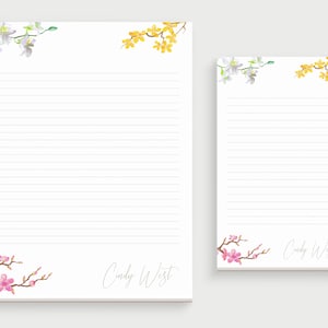 May include: Two lined notepad sets with floral designs. The larger notepad has white and yellow flowers in the top corners and pink flowers in the bottom corners. The smaller notepad has white and yellow flowers in the top corners. Both notepads have the name "Cindy West" written at the bottom.