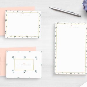 May include: Two white stationery cards with a gold border and a green floral pattern. The top card has the name "Anne Marie Doulin" printed in pink. The bottom card has the same name printed in pink with a gold border.