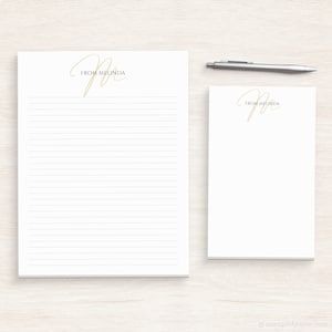 May include: Two white notepad sets with lined paper. The top of each notepad has a gold cursive "M" with the text "From Melinda" underneath.