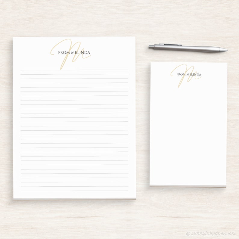 May include: Two white notepad sets with lined paper. The top of each notepad has a gold cursive "M" with the text "From Melinda" underneath.