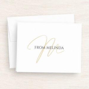 May include: A white card with a gold cursive letter "M" and the text "FROM MELINDA" in black.