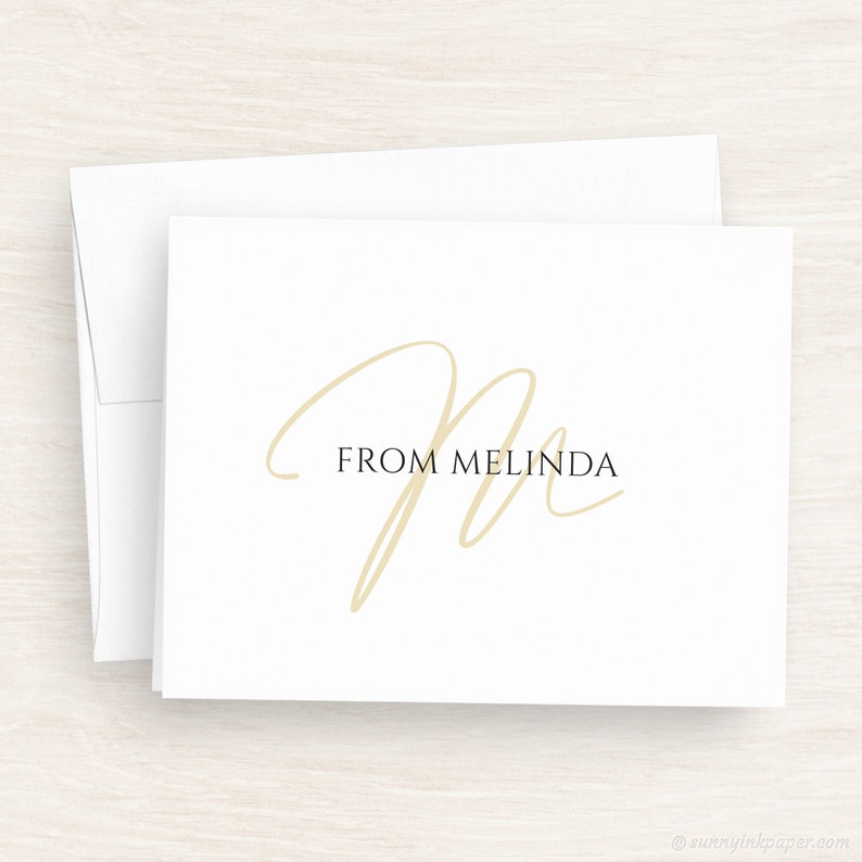 May include: A white card with a gold cursive letter "M" and the text "FROM MELINDA" in black.