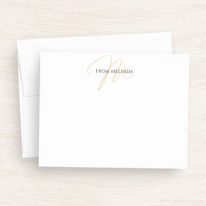 May include: A white card with a gold cursive letter "M" and the text "From Melinda" printed in black.