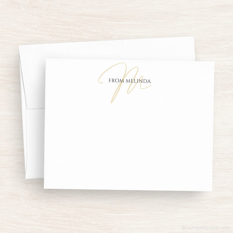 May include: A white card with a gold cursive letter "M" and the text "From Melinda" printed in black.