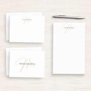 Op de afbeelding: Twee sets wit briefpapier met een gouden cursief "M" en de tekst "From Melinda".