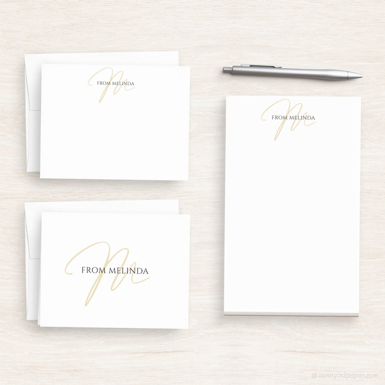 Op de afbeelding: Twee sets wit briefpapier met een gouden cursief "M" en de tekst "From Melinda".