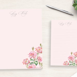 May include: Two pink personalized stationery notepads with a floral design. The top notepad has the name "Lucy Cole" written in cursive. The bottom notepad has the same name and a floral design in the bottom right corner.