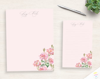 May include: Two pink personalized stationery notepads with a floral design. The top notepad has the name "Lucy Cole" written in cursive. The bottom notepad has the same name and a floral design in the bottom right corner.