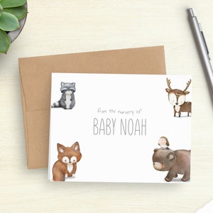 May include: A white baby announcement card with watercolor illustrations of woodland animals, including a raccoon, fox, deer, and bear. The text "from the nursery of BABY NOAH" is centered on the card. A brown envelope and a silver pen are also in the image.