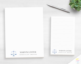 May include: A white stationery set featuring a blank sheet and a lined notepad. Both items display a blue scale of justice icon, the name MARVIN LESTER, the title ATTORNEY AT LAW, and the phone number (800) 765-4321 printed in black ink.