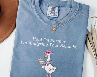 May include: A blue t-shirt with a graphic of a white goose wearing a pink cowboy hat, pink boots, and a pink bandana. The goose holds a clipboard and pencil. The text reads: Hold On Partner, I'm Analyzing Your Behavior. Includes a gold necklace.