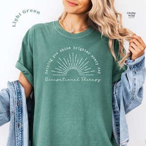 May include: Light green t-shirt with the text "Helping you shine brighter every day" and "Occupational Therapy" in white. The shirt has a crew neck and short sleeves. The shirt is made of a soft material.