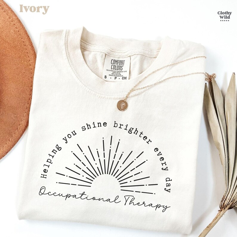 May include: Ivory-colored t-shirt with the text "Helping you shine brighter every day Occupational Therapy" in black. A gold necklace with a pendant rests on the shirt. The shirt is from Comfort Colors.