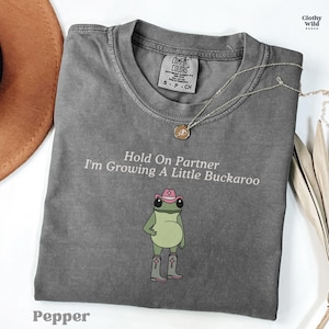 May include: Pepper-colored t-shirt with the text "Hold On Partner I'm Growing A Little Buckaroo" and a cartoon frog wearing a cowboy hat and boots. A silver necklace and a brown hat are also visible.