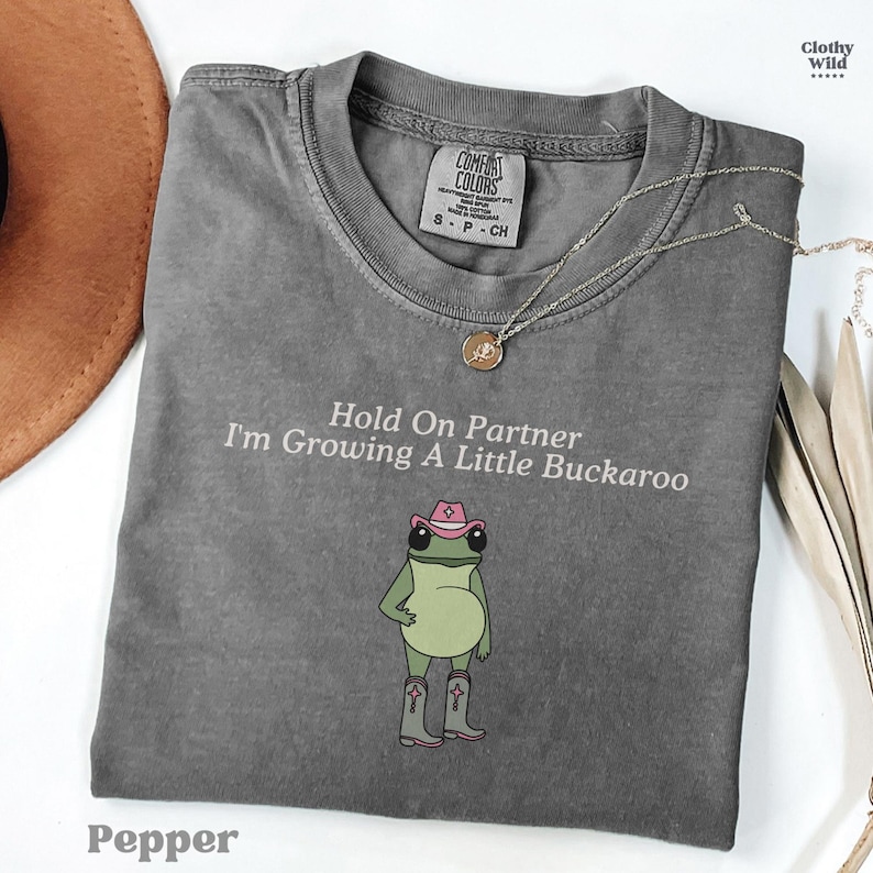 May include: Pepper-colored t-shirt with the text "Hold On Partner I'm Growing A Little Buckaroo" and a cartoon frog wearing a cowboy hat and boots. A silver necklace and a brown hat are also visible.