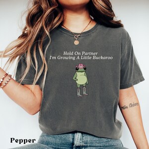 May include: Pepper-colored t-shirt with the text "Hold On Partner I'm Growing A Little Buckaroo" and a cartoon frog wearing a pink cowboy hat and boots. The shirt is a relaxed fit.
