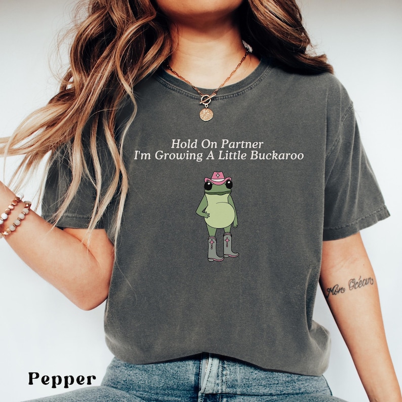 May include: Pepper-colored t-shirt with the text "Hold On Partner I'm Growing A Little Buckaroo" and a cartoon frog wearing a pink cowboy hat and boots. The shirt is a relaxed fit.
