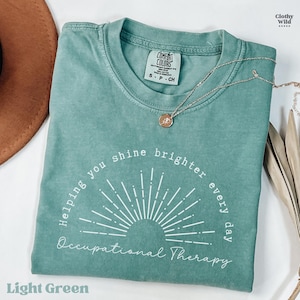 May include: Light green t-shirt with the text "Helping you shine brighter every day Occupational Therapy" in white. The shirt has a sunburst design and a Comfort Colors tag. A silver necklace with a pendant is on the shirt.