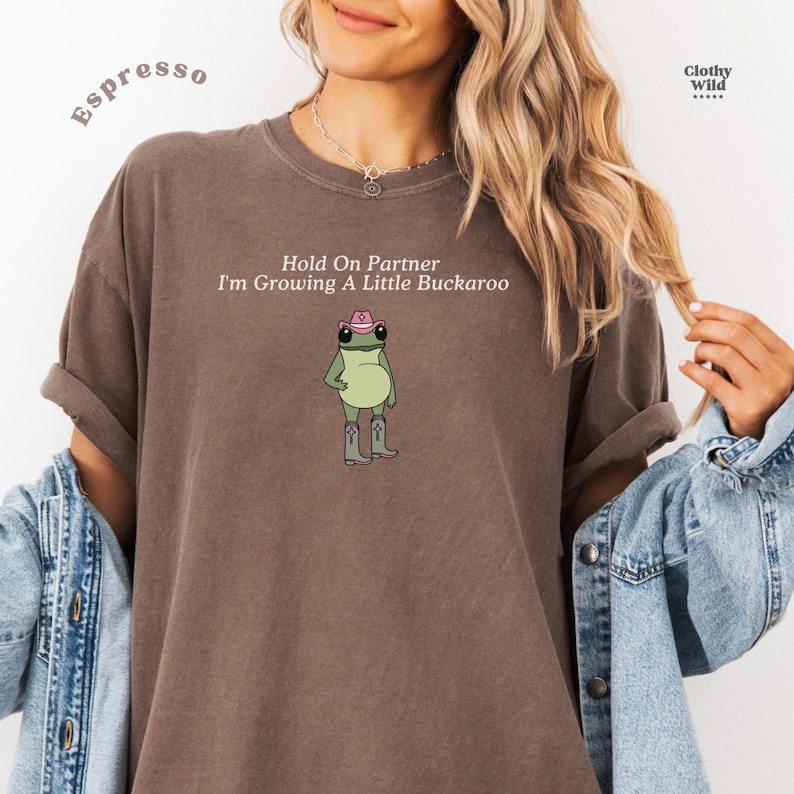 May include: Espresso-colored t-shirt with the text "Hold On Partner I'm Growing A Little Buckaroo." The shirt features a cartoon frog wearing a cowboy hat and boots. The shirt is from Clothy Wild.