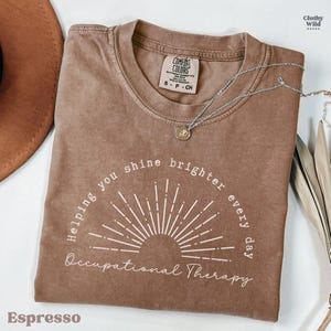 May include: Espresso-colored t-shirt with the text "Helping you shine brighter every day Occupational Therapy" in white. The shirt has a crew neck and short sleeves. A silver necklace with a pendant is draped on the shirt.