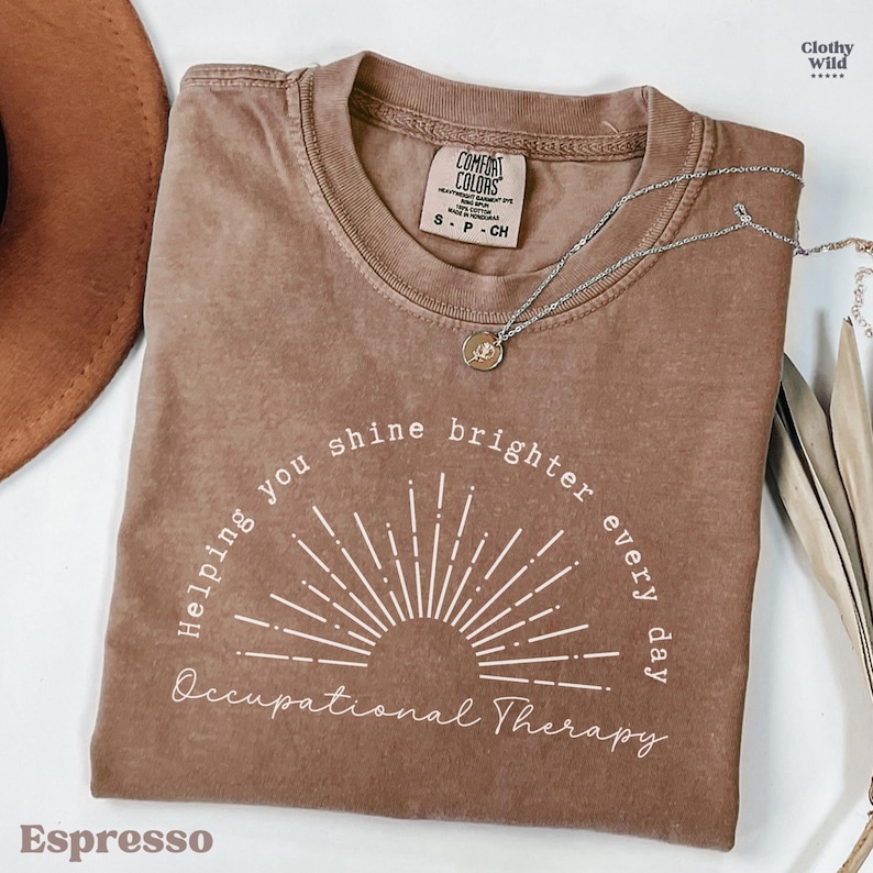 May include: Espresso-colored t-shirt with the text "Helping you shine brighter every day Occupational Therapy" in white. The shirt has a crew neck and short sleeves. A silver necklace with a pendant is draped on the shirt.