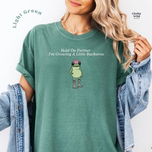 May include: Light green t-shirt with the text "Hold On Partner I'm Growing A Little Buckaroo" and a cartoon frog wearing a cowboy hat and boots. The shirt has rolled-up sleeves and a relaxed fit.