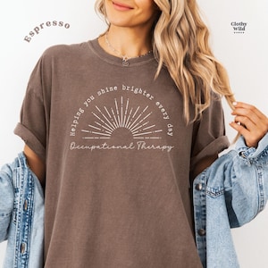 May include: Espresso-colored t-shirt with the text "Helping you shine brighter every day" and "Occupational Therapy" in a white, stylized font. The design includes a sunburst graphic. The shirt is a relaxed fit.