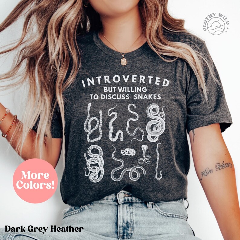 Puede incluir: Camiseta de color gris oscuro jaspeado con texto blanco que dice "Introvertido pero dispuesto a hablar de serpientes". La camiseta presenta un gr&aacute;fico de varias serpientes en estilo vintage.
