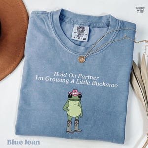 May include: Blue jean colored t-shirt with the text "Hold On Partner I'm Growing A Little Buckaroo". The shirt features a cartoon frog wearing a pink cowboy hat and boots. A gold necklace is draped across the shirt.