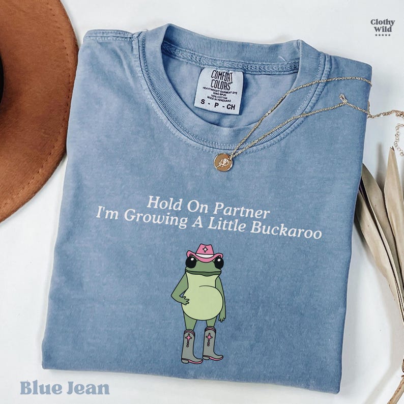 May include: Blue jean colored t-shirt with the text "Hold On Partner I'm Growing A Little Buckaroo". The shirt features a cartoon frog wearing a pink cowboy hat and boots. A gold necklace is draped across the shirt.