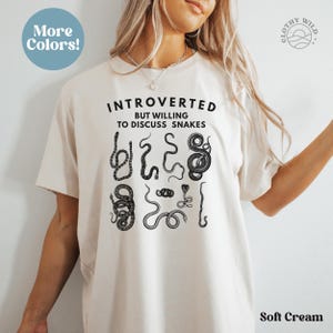Puede incluir: Una camiseta de color crema suave con el texto "Introvertido pero dispuesto a hablar de serpientes" y un gr&aacute;fico de varias serpientes. El texto "&iexcl;M&aacute;s colores!" est&aacute; en un c&iacute;rculo en la esquina superior izquierda. El texto "Clothy Wild" est&aacute; en un c&iacute;rculo en la esquina superior derecha.