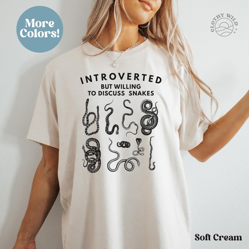 Puede incluir: Una camiseta de color crema suave con el texto "Introvertido pero dispuesto a hablar de serpientes" y un gr&aacute;fico de varias serpientes. El texto "&iexcl;M&aacute;s colores!" est&aacute; en un c&iacute;rculo en la esquina superior izquierda. El texto "Clothy Wild" est&aacute; en un c&iacute;rculo en la esquina superior derecha.