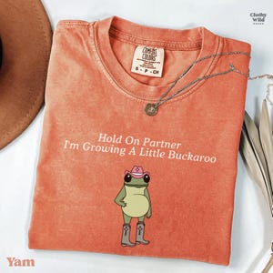 May include: A coral Comfort Colors t-shirt with the text "Hold On Partner I'm Growing A Little Buckaroo" and a cartoon frog wearing a pink cowboy hat and boots. A silver necklace with a pendant is draped on the shirt.