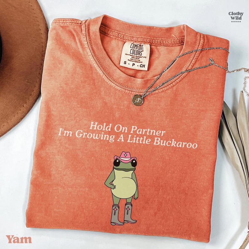 May include: A coral Comfort Colors t-shirt with the text "Hold On Partner I'm Growing A Little Buckaroo" and a cartoon frog wearing a pink cowboy hat and boots. A silver necklace with a pendant is draped on the shirt.