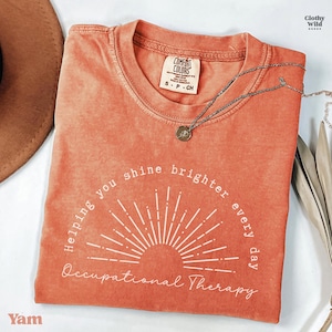 May include: A coral-colored t-shirt with the text "Helping you shine brighter every day Occupational Therapy" in white. A silver necklace with a pendant rests on the shirt. The shirt is a Comfort Colors brand, size S-P-CH.