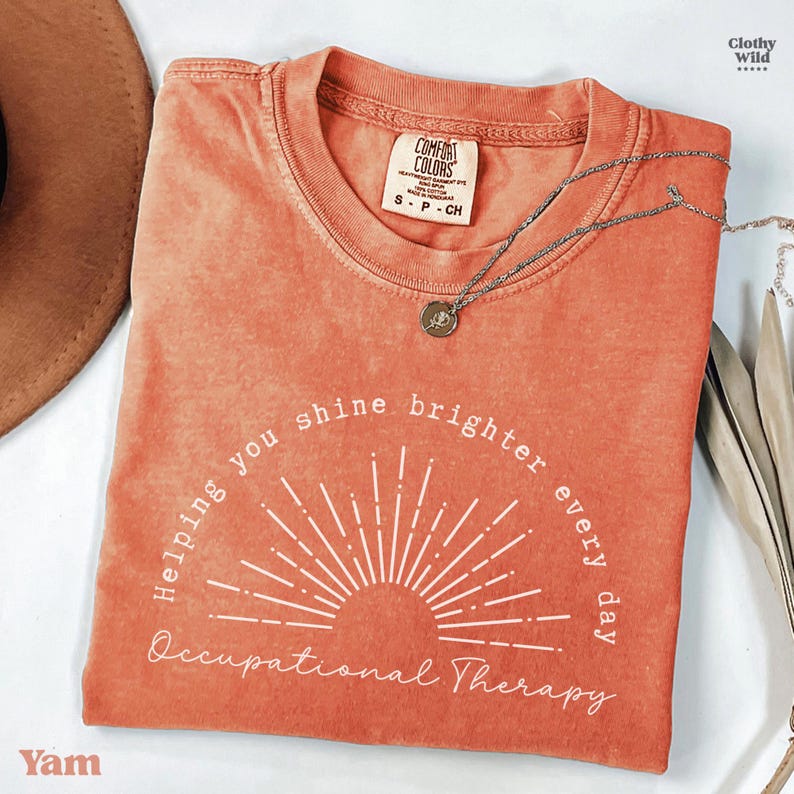 May include: A coral-colored t-shirt with the text "Helping you shine brighter every day Occupational Therapy" in white. A silver necklace with a pendant rests on the shirt. The shirt is a Comfort Colors brand, size S-P-CH.