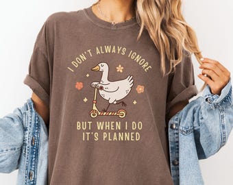 May include: A dark brown short-sleeve t-shirt featuring a graphic of a white goose riding a scooter. The text reads, I DON'T ALWAYS IGNORE BUT WHEN I DO IT'S PLANNED. The design includes small orange flowers and star shapes around the goose.