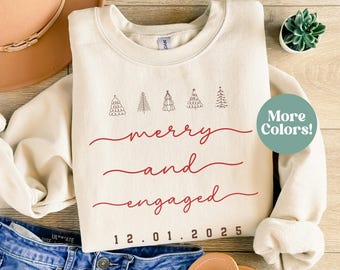 May include: A sand-colored sweatshirt with the words "merry and engaged" in red script, along with the date "12.01.2025". Above the text are five small tree illustrations. A casual, festive design.
