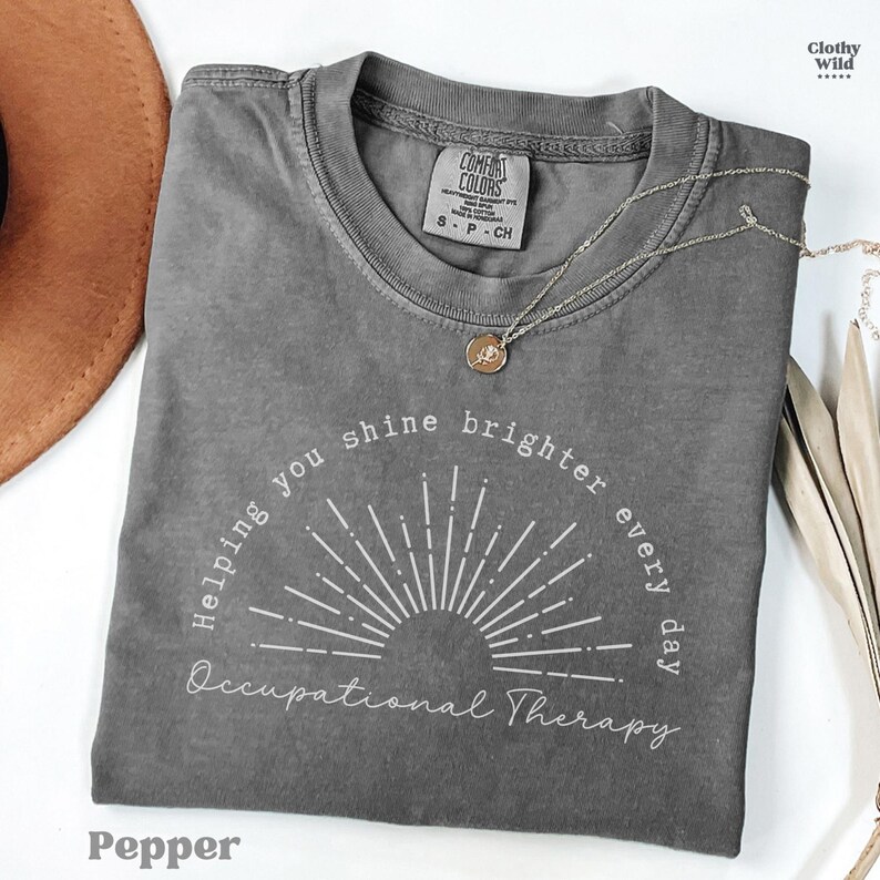 May include: A gray Comfort Colors t-shirt with the text "Helping you shine brighter every day Occupational Therapy." The shirt has a crew neck and short sleeves. A silver necklace with a pendant is draped across the shirt. The word "Pepper" is at the bottom.