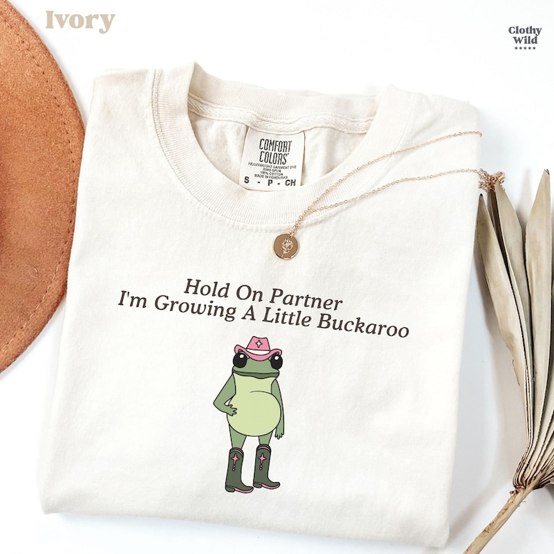 May include: Ivory-colored t-shirt with the text "Hold On Partner I'm Growing A Little Buckaroo" and a cartoon frog wearing a cowboy hat and boots. A gold necklace is draped across the shirt. The shirt is from Comfort Colors.