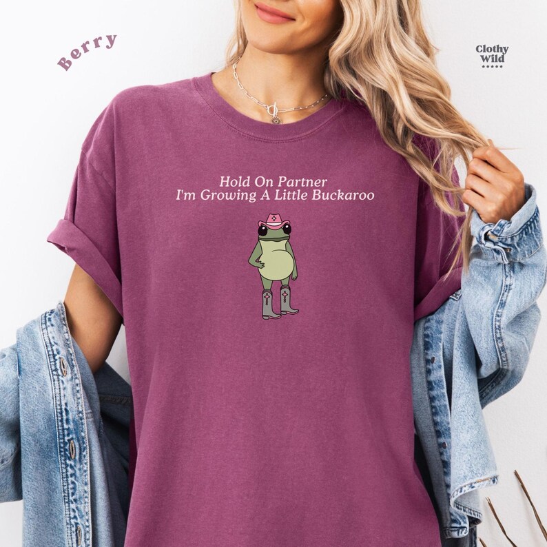 May include: Berry-colored t-shirt with the text "Hold On Partner I'm Growing A Little Buckaroo" and a cartoon frog wearing a cowboy hat and boots. The shirt has rolled-up sleeves and a crew neck.